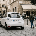 Un’auto elettrica bianca transita su una strada lastricata in un centro storico, passando accanto a una donna a piedi. Sullo sfondo si vede un bar con persone sedute ai tavoli.