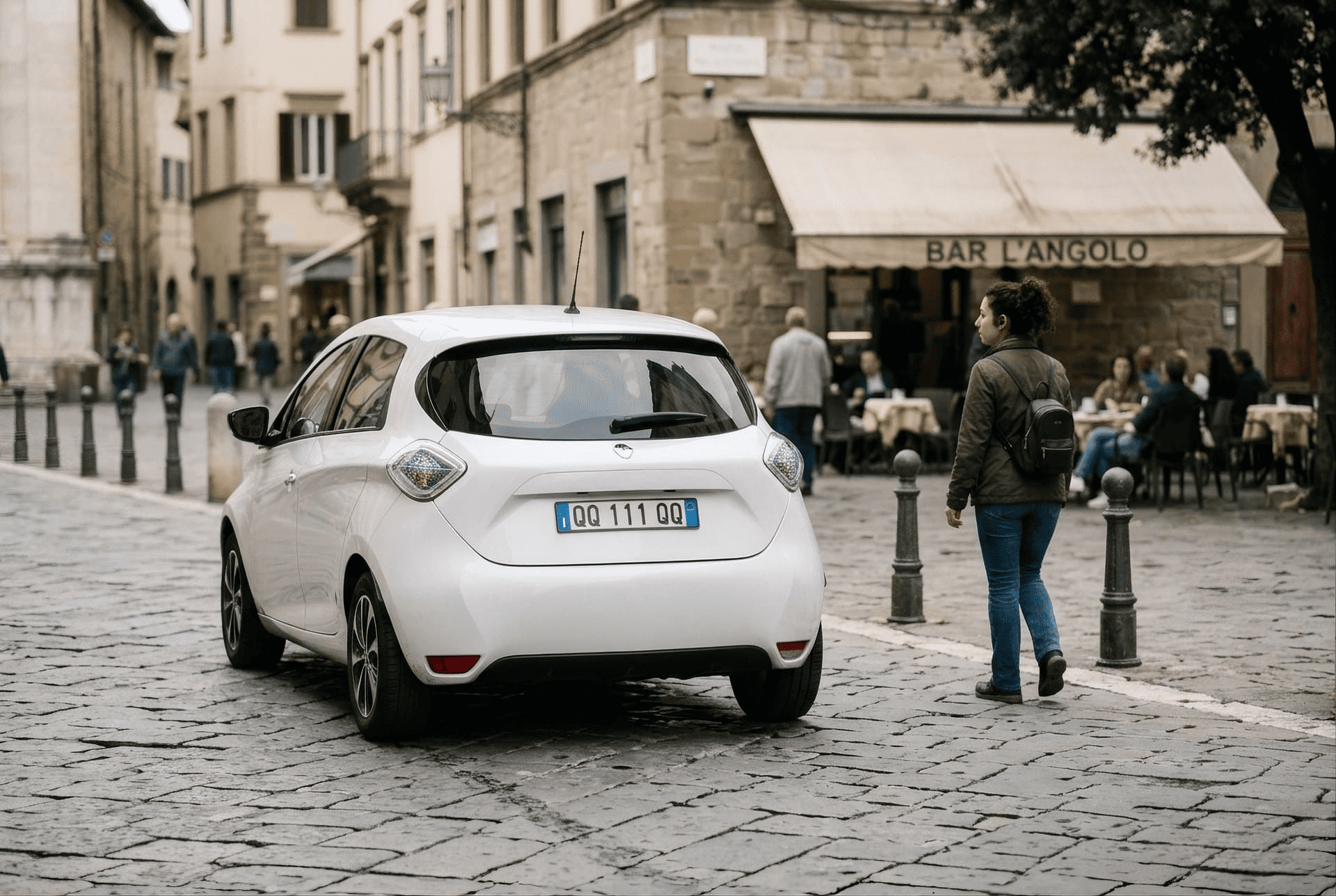 Un’auto elettrica bianca transita su una strada lastricata in un centro storico, passando accanto a una donna a piedi. Sullo sfondo si vede un bar con persone sedute ai tavoli.